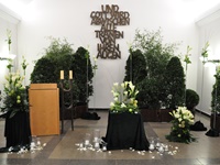 Feierhalle mit Dekorationsbäumen, 2 Gestecke in weiß, Dekoration mit Lilien, Urnendekoration, Kerzen und Rosenblütenblätter